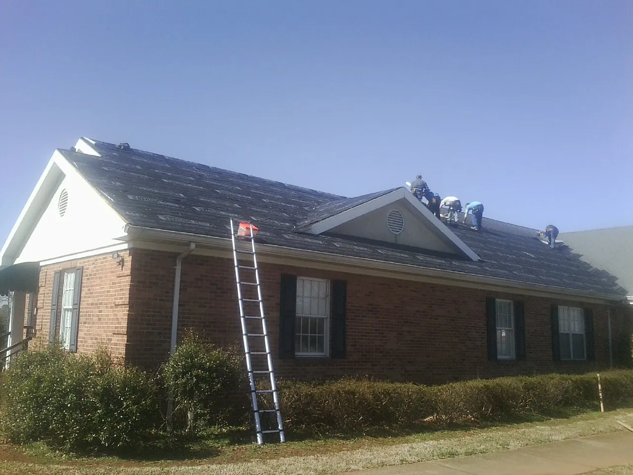 Crew tearing shingles off a brick ranch roof with a ladder set — Emergency Roof Repair work by our crew in La Vernia