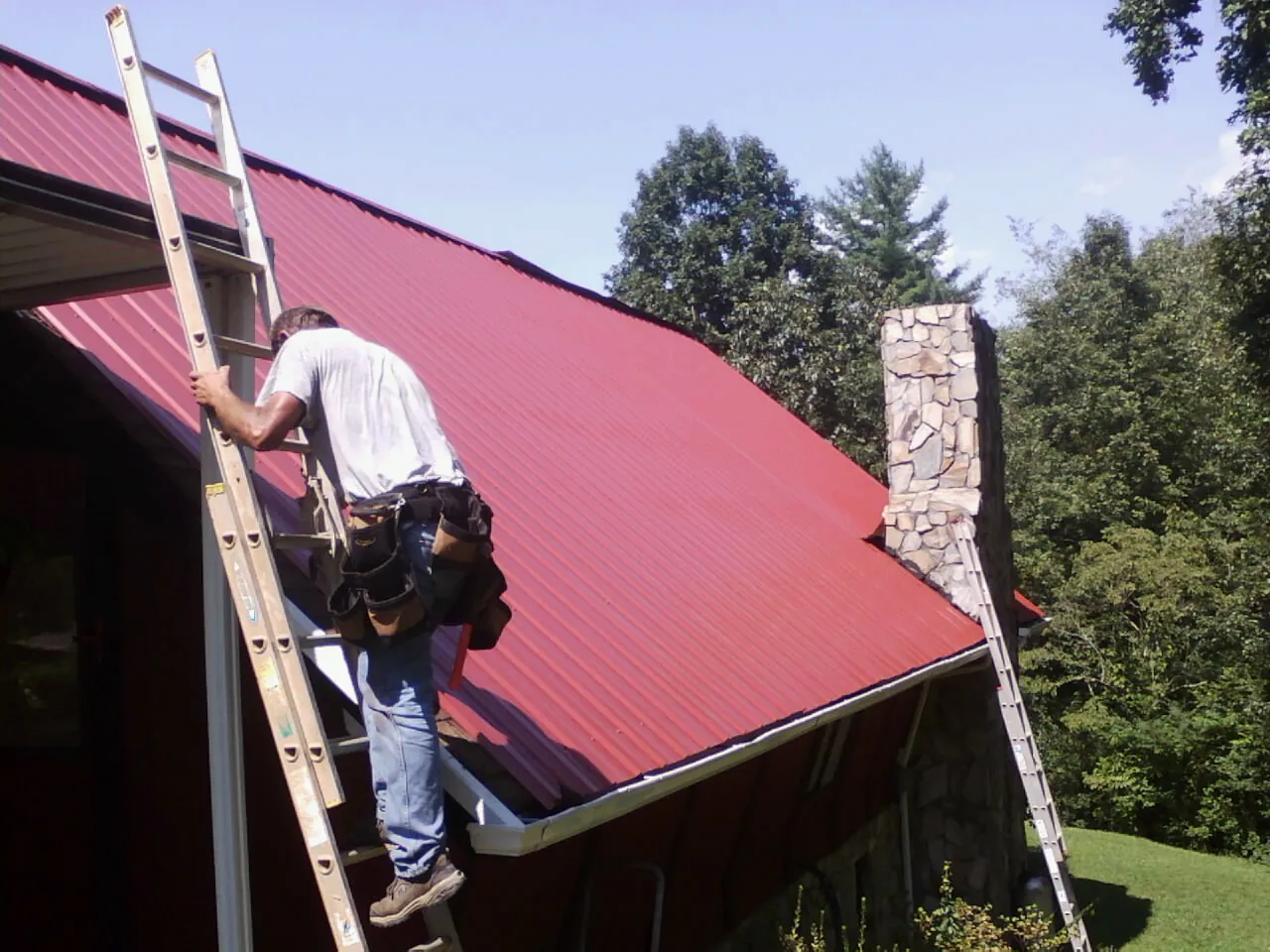 Roofer on a ladder beside a red metal roof and stone chimney — Soffit & Fascia Repair by Roofing Brothers in La Vernia